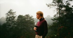 Woman in a burgundy turtleneck holding a glass of red wine on a misty forested terrace